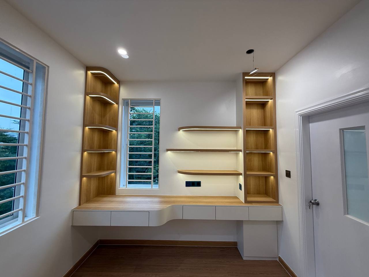 Built-in desk between two illuminated wood-toned shelves beside windows